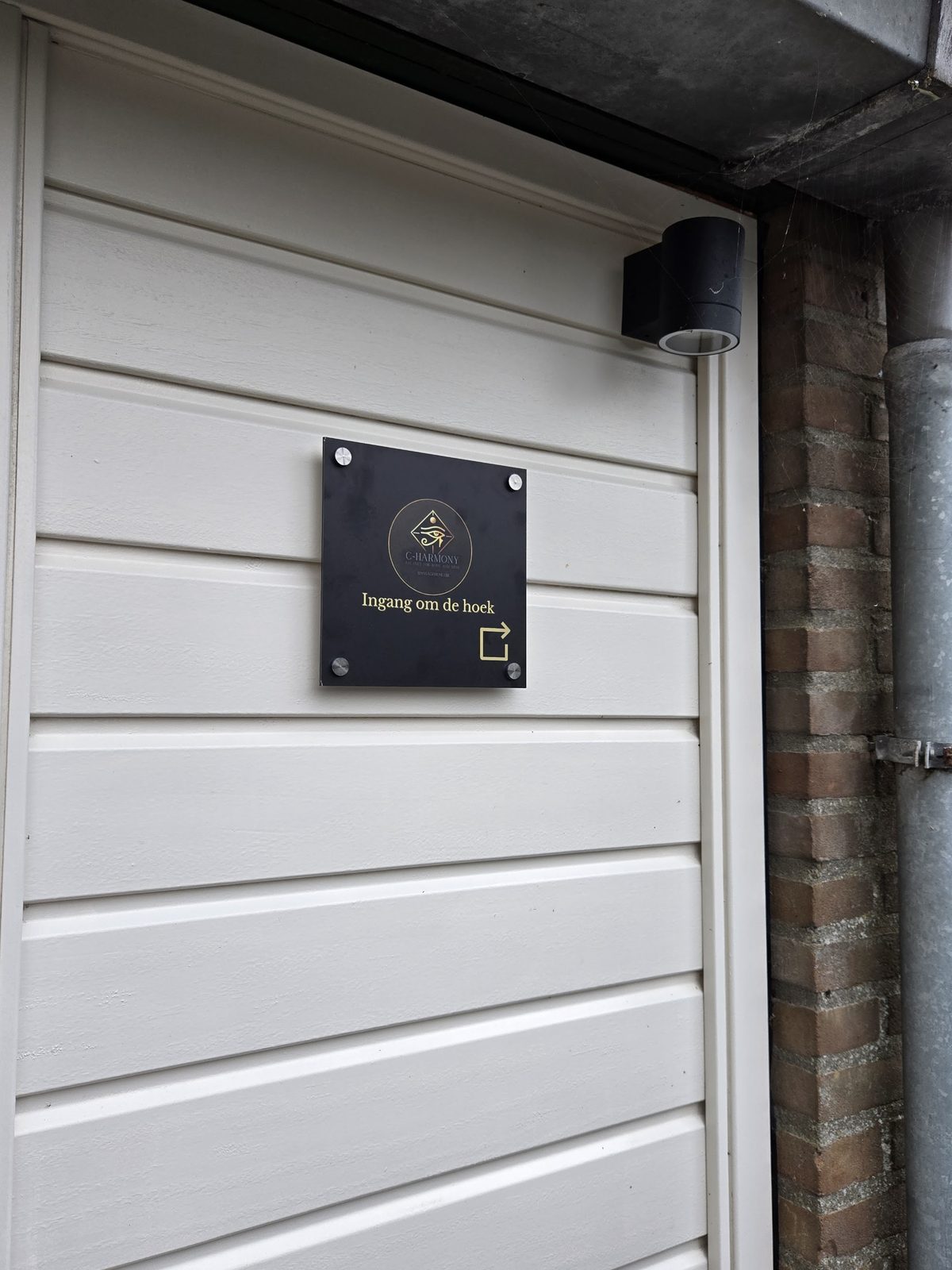 Zwart bordje met het gouden C-harmony logo en de tekst Ingang om de hoek